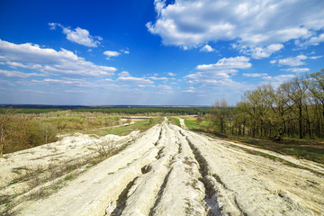 Beautiful view of road in the field and green forest and blue cloudy sky from the top of the hill. Spring rural landscape.