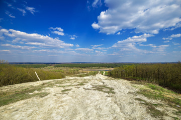 Beautiful view of road in the field and green forest and blue cloudy sky from the top of the hill. Spring rural landscape.