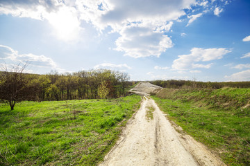Beautiful view of road in the field and green forest and blue cloudy sky and top of the hill. Spring rural landscape.
