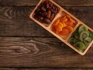 Dates, dried apricots and kiwis in a Compartmental dish on a dark wooden table.
