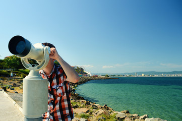 A young man on vacation looks through a white telescope against a landscape with sea and mountains in the Bulgarian town of Nessebar