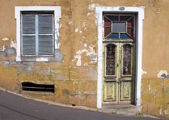 Fototapeta na wymiar the front of an old abandoned yellow house with blue shuttered windows and locked green wooden door with flaking peeling paint on a sloping street