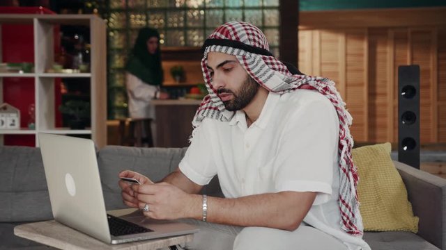 Handsome arabic muslim man in traditional costume using credit card as payment method when shopping online using laptop. Technologies concept. Being online, surfing internet
