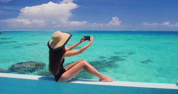 Phone - woman taking photo pictures using mobile cell phone app on vacation by pool by tropical ocean coral lagoon on travel holidays. Slow motion video shot on RED Cinema Camera.