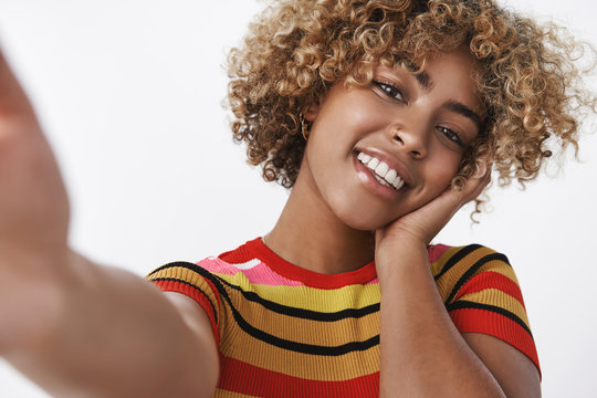 Selfie shot of stylish african american woman with pierced nose and fair curly haircut tilting head carefree and tender touching cheek gently holding camera with extended hand over white background