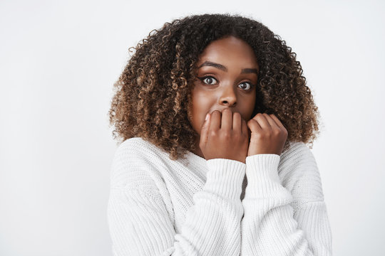 Waist-up shot of intense scared cute african-american woman watching scary movie gasping and jumping from frightening sound holding hands on lips biting fingernails nervously looking at camera worried