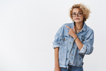 Looks suspicious what you think. Portrait of doubtful unsure and hesitant good-looking stylish girlfriend in denim jacket and glasses pursing chin in judgemental smirk pointing at upper left corner