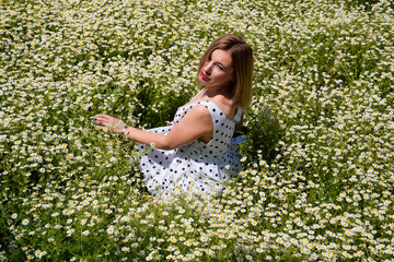 Naklejka na ściany i meble A woman in a white dress with polka dots is on a glade with daisies. Blooming daisies in the field.