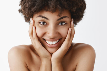 Attractive feminine and natural african american young female with curly hair and pure clean skin, touching face and smiling broadly with excitement and happiness, posing naked over grey wall
