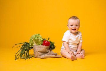 baby girl sitting with bag of vegetables on yellow isolate background, space for text. the concept of healthy eating.organic food