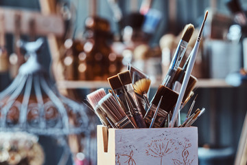 Jar with different brushes on the table at art studio. There are decorative cage at background.
