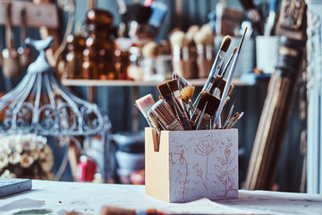 Jar with different brushes on the table at art studio. There are decorative cage at background.