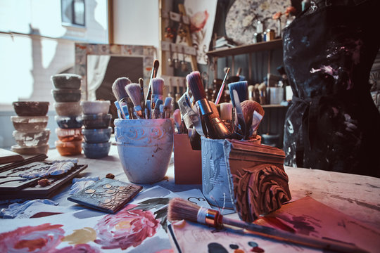 There are dark and a lot of different brushes on artist's table in jars. There are artistick's mess on the table.