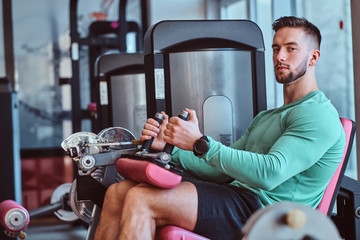 Strong pensive man is sitting on training apparatus in gym and doing legs exercises.