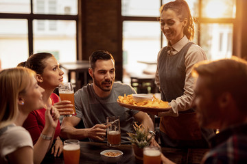Happy waitress serving nacho chips to group of friend in a pub.