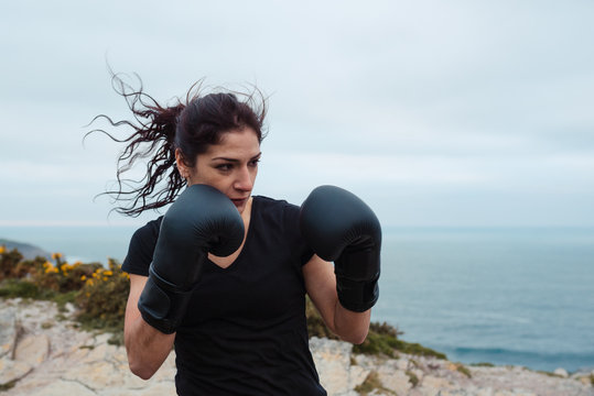 Man and woman in boxing gloves punching each other while standing on cliff against sea and cloudless sky