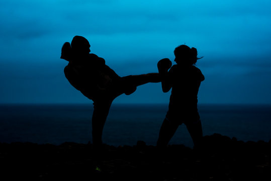 Silhouettes of anonymous fighters practicing kickboxing against sea and dark sky in evening in countryside