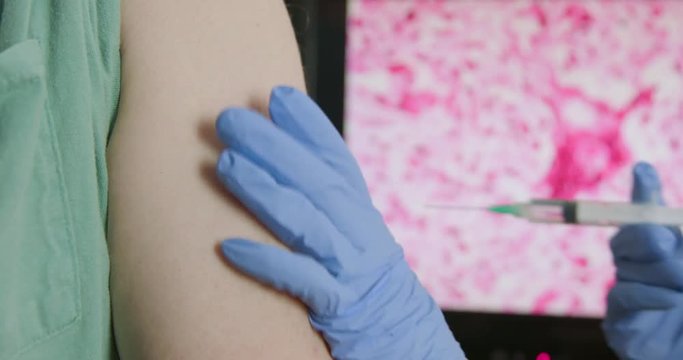 A doctor or nurse preparing to vaccinate someone against the measles using a needle and syringe with the virus displayed on a digital monitor in the background.