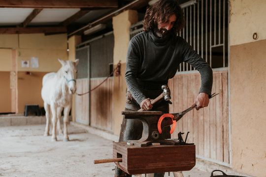 Adult farrier using hammer and tongs to forge hot horseshoe on portable anvil near stable on ranch