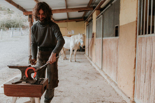 Adult farrier using hammer and tongs to forge hot horseshoe on portable anvil near stable on ranch