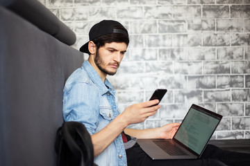 Young guy sitting on the floor, looking in smartphone with laptop on legs. Wearing black cap and blue jeans jacket.