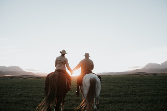 Back view of man and woman riding horses and holding hands against sunset sky on ranch