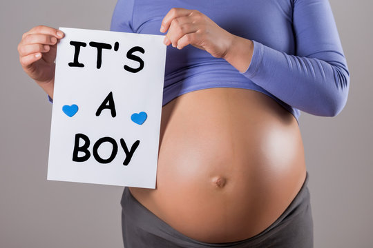 Image of close up stomach of pregnant woman holding paper with text it's a boy on gray background.
