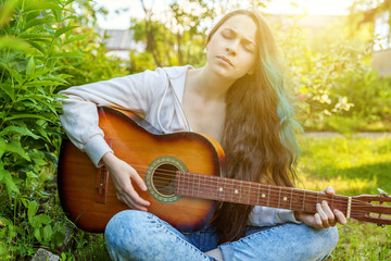 Young hipster woman sitting in grass and playing guitar on park or garden background. Teen girl...