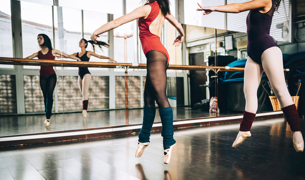 Crop women in bodysuits jumping on pointe above floor in studio training ballet.