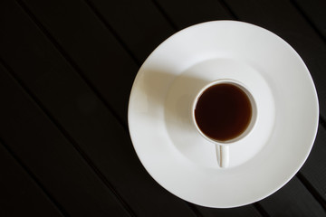 A white cup of black tea stands in a saucer on a dark wooden table. View from above. Minimalism
