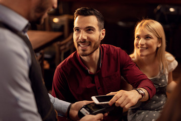 Happy man making contactless payment via smart phone in a cafe.