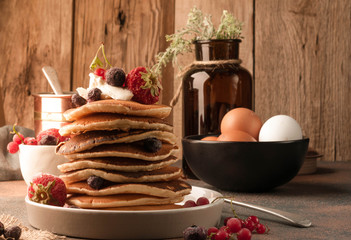 traditional american fried stacked pancakes with fresh berries in front of bowl with eggs and glass bottle and herbs on table