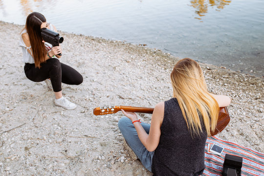 Young lady in casual outfit using camera to shoot anonymous female friend playing acoustic guitar while spending time on beach together