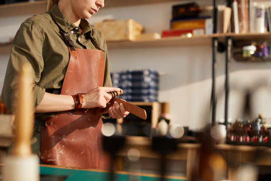 Mid section portrait of young woman working with leather while making handcrafted belt in shop lit by sunlight, copy space