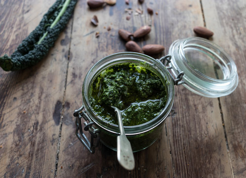 Closeup glass jar with yummy kale pesto placed on lumber tabletop near fresh peanuts