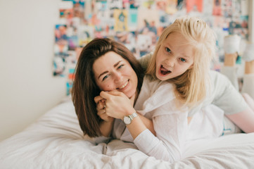 Obraz na płótnie Canvas Lifestyle soft focus portrait of happy mom hugs her adorable daughter on white bed. Indoor happy family portrait of smiling mother and her daughter lying together on bed with decorative wall behind.
