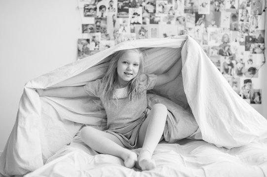 Funny little girl sitting on white bed and holding blanket above her head. Lifestyle black white portrait of happy smiling blonde female kid covering herself with blanket on decorative wall background
