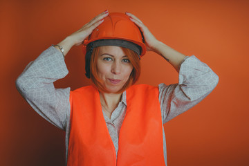 Adult female with red hair in working uniform on orange background. Portrait of woman in bright orange protective hardhat and vest looking at camera on orange background