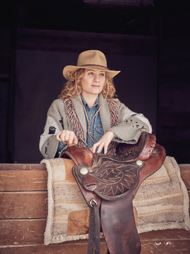 Beautiful cowgirl looking away while leaning on wooden stable wall near leather saddle on ranch