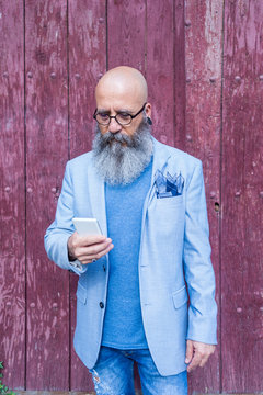 Front view of a mature trendy bearded hipster standing against a red vintage door outdoors while using a mobile phone
