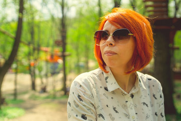 Young woman with red hair, sunglasses, among the trees. Portrait of young attractive woman with fashionable hairstyle sitting in Park