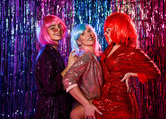 Three friends in wigs and stylish glamorous dresses with sequins, in the neon light of a disco. Celebrate, have fun, celebrate something