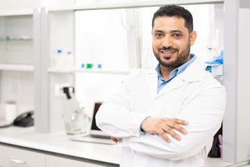 Smiling handsome young middle-eastern lab worker with beard in white coat crossing arms on chest...
