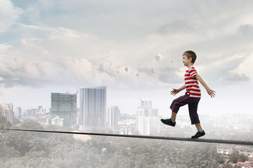 Cute joyful little boy walking on a rope over city. Mixed media