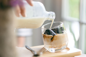 Closeup image of a woman pouring milk into a glass of ice cube coffee in cafe