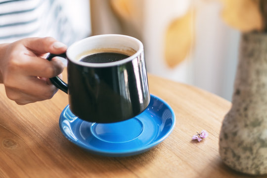 Closeup image of a woman's hand holding a black cup of hot coffee on wooden table