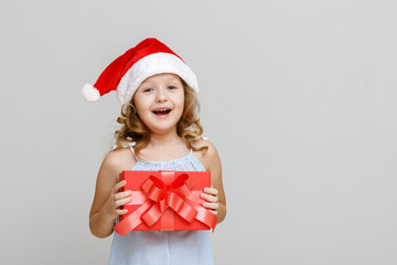 Portrait of a happy smiling little blonde girl on a gray background. A child in a Santa hat is holding a red crust with a gift. Christmas celebration concept