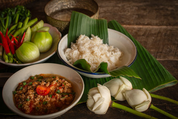 Thai Northern Style Pork and Tomato Chili Relish, nam prik ong in white bowl on wood table there are side dishes of fresh vegetables, cooked rice, water bowl and flower placed around.