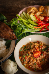 Thai Northern Style Pork and Tomato Chili Relish, nam prik ong in white bowl on wood table there are side dishes of fresh vegetables, cooked rice and flower placed around.