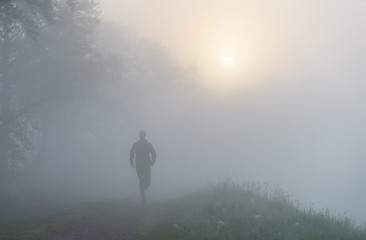 Naklejka na ściany i meble Athlete running on a gravel road during a foggy, spring sunrise in the countryside.